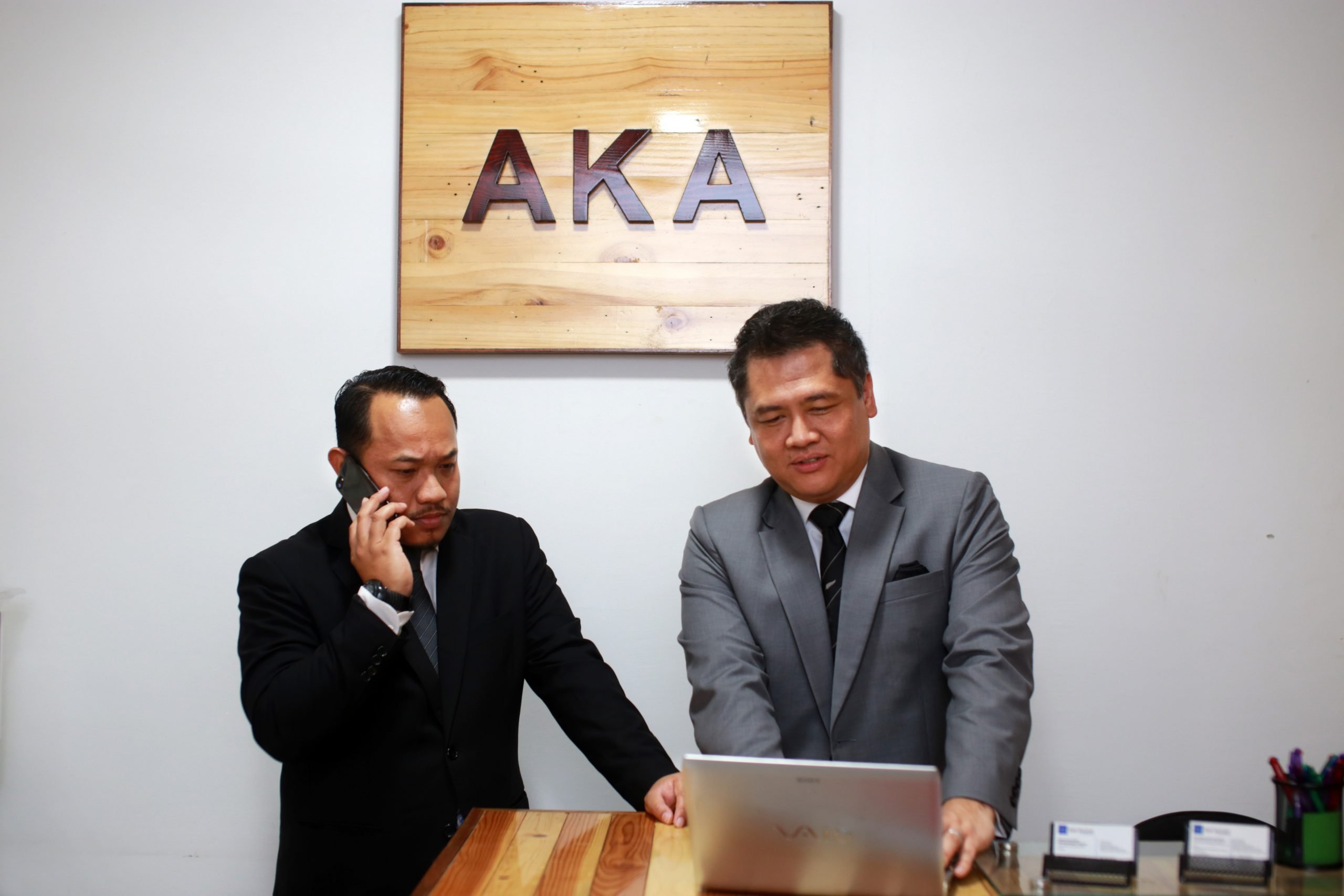 Two formally dressed men having a discussion in an office, one is making a call while the other is looking at the laptop's screen.