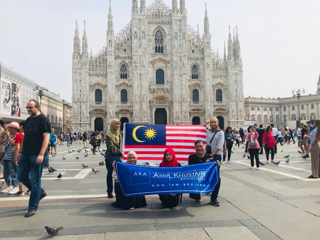 Five people posing for a picture in front of a tourist attraction, holding the Malaysia's flag and a blue colored banner of Amir Khusyairi & Associate Legal Firm.
